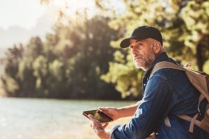Senior man at a lake with backpack and digital tablet